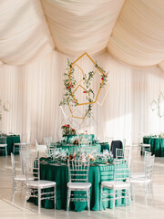 The decor of the wedding banquet hall with tables in emerald green, white drapery on the ceiling, and gold geometric decorative elements.