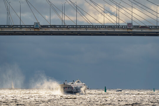 a high-speed passenger hydrofoil boat passes under a cable-stayed bridge in sunny weather, dark storm clouds in the background