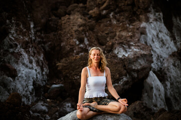 beautiful woman sitting in meditation against dark rocks