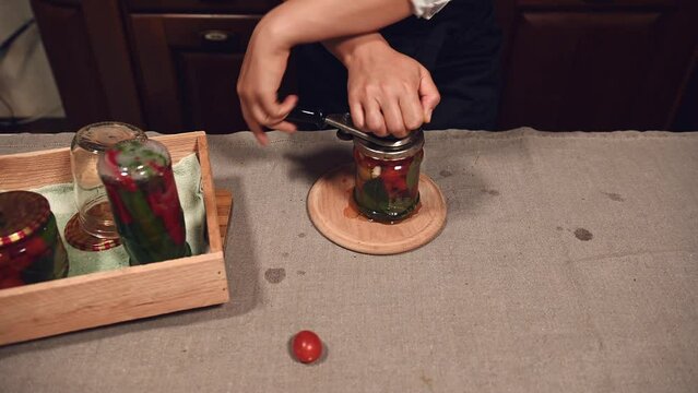 Steadicam shot. View from above of a housewife closes lids on the jars with canned cherry tomatoes, with a seamer or special seaming key. Canning. Pickling. Cooking homemade tinned food. Marinating