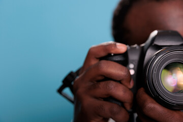 Close up shot of young african american photographer taking photo with professional DSLR camera while standing on blue background. Photography enthusiast with modern photo device taking picture.