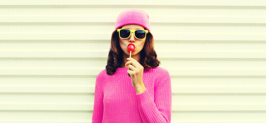 Portrait of stylish young woman with lollipop wearing pink hat, sweater on white background