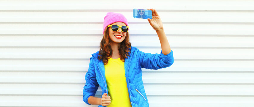 Portrait of modern happy smiling young woman taking selfie with smartphone wearing colorful pink hat, blue jacket on white background