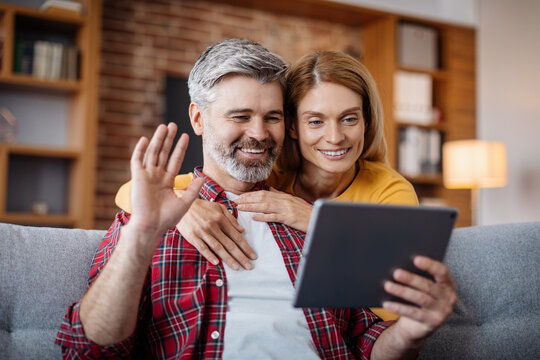 Cheerful mature caucasian female hugging man, waving hand, watching video in tablet, calling online