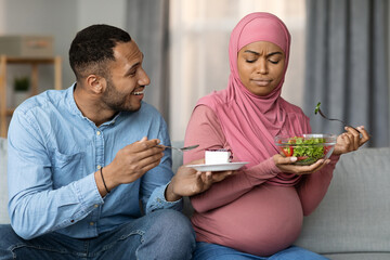 Husband Offering Cake To Pregnant Muslim Wife, Woman Frowning And Eating Salad