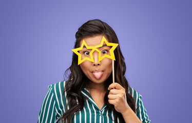 party props, photo booth and people concept - happy woman with big glasses in shape of stars sticking out her tongue over violet background