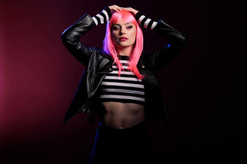 Attractive trendy girl with pink hair wearing leather jacket in studio with dark light, posing over background. Confident woman with stylish rocker clothes and perfect makeup look, fashion style.