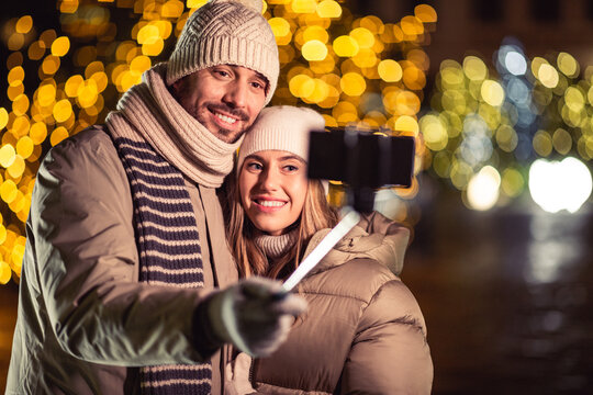 winter holidays and people concept - happy smiling couple taking picture with smartphone on selfie stick and hugging over christmas lights in evening