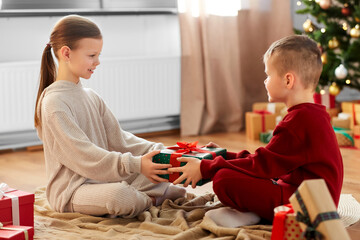 christmas, winter holidays and childhood concept - happy girl and boy in pajamas holding gift box together sitting on floor in front of each other at home