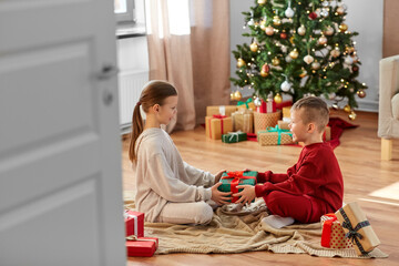 Naklejka premium christmas, winter holidays and childhood concept - happy girl and boy in pajamas holding gift box together sitting on floor in front of each other at home