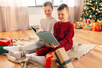 christmas, winter holidays and childhood concept - happy girl and boy in pajamas reading book...