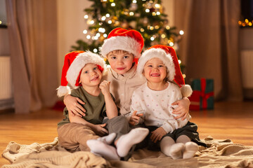 Fototapeta na wymiar christmas, winter holidays and childhood concept - happy little girl and boys in santa hats hugging on floor at home