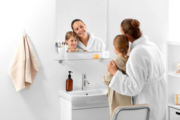 Obraz premium beauty, family and people concept - happy smiling mother and daughter with moisturizer on their faces looking to mirror in bathroom