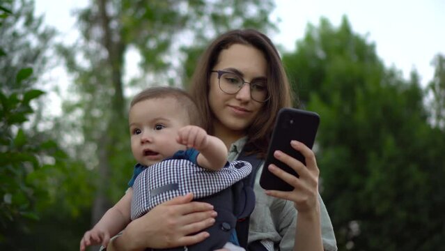 A young woman with a child stands with a phone in her hands in the park. Mom in glasses with her son in her arms. Newborn son in a kangaroo-backpack. Close-up.
