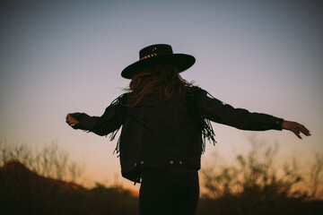 Woman in hat and fringe jacket dancing at sunset with back turned