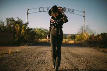 Woman posing in front of a ranch in western fashion and cowboy hat - obrazy, fototapety, plakaty