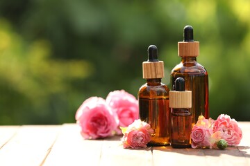 Bottles of rose essential oil and flowers on wooden table outdoors, space for text