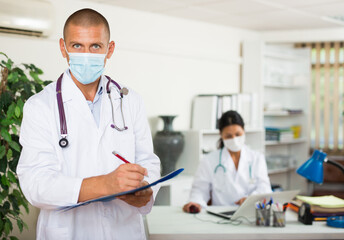 Fototapeta Focused therapist in white coat and protective face mask standing in clinic, filling clipboard with medical records obraz