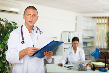 Fototapeta na wymiar Positive male doctor standing in modern office with documents, writing medical history sheet
