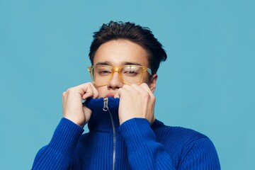 a cute, handsome man stands on a light blue background in a blue zip-up sweater with yellow sunglasses on his face and looks at the camera, turning up his collar and hiding his face