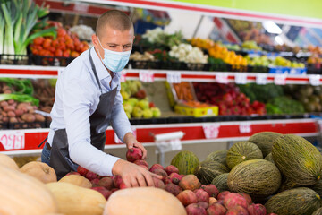 Salesman wearing protective mask to prevent viral infection working in greengrocery, arranging fruits and vegetables on shelves