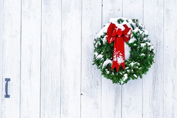 Snow-dusted pine needle Christmas wreath and red bow on weathered, whitewashed wide plank door...