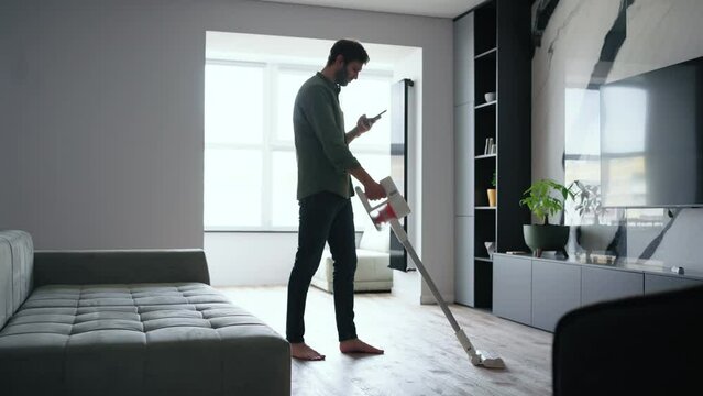 A man with a mobile phone in his hands reads social media and does routine chores around the house with a wireless vacuum cleaner