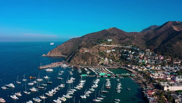 Very high drone shot flying over the harbor of Avalon on Catalina Island with a view of sail boats, palm trees, and the Wrigley mansion hotel.