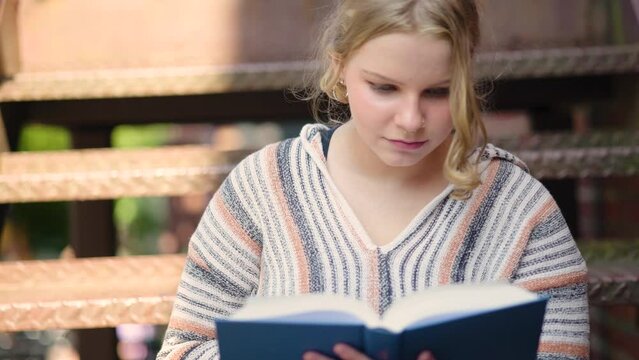 Close up of a girl student turning a page while reading a book outside on stairs. Slow motion.