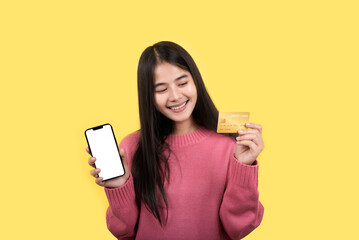Portrait of a delighted woman standing on a yellow background. holding a cell phone showing a plastic credit card - obrazy, fototapety, plakaty