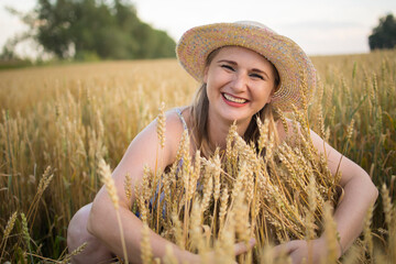 A beautiful middle-aged farmer woman in a straw hat and a plaid shirt stands in a field of golden ripening wheat during the daytime in the sunlight