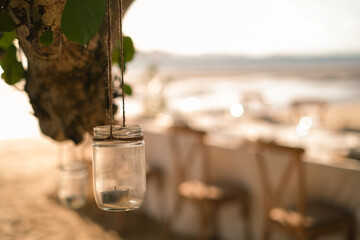 Close up the jar candle hanging on the tree with long table wedding dinner setup on the beach at Thailand in the evening. Wedding party concept. Decoration outdoor restaurant at the beach.