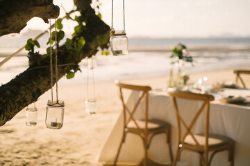 Close up the jar candle hanging on the tree with long table wedding dinner setup on the beach at Thailand in the evening. Wedding party concept. Decoration outdoor restaurant at the beach.
