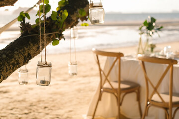 Close up the jar candle hanging on the tree with long table wedding dinner setup on the beach at Thailand in the evening. Wedding party concept. Decoration outdoor restaurant at the beach.