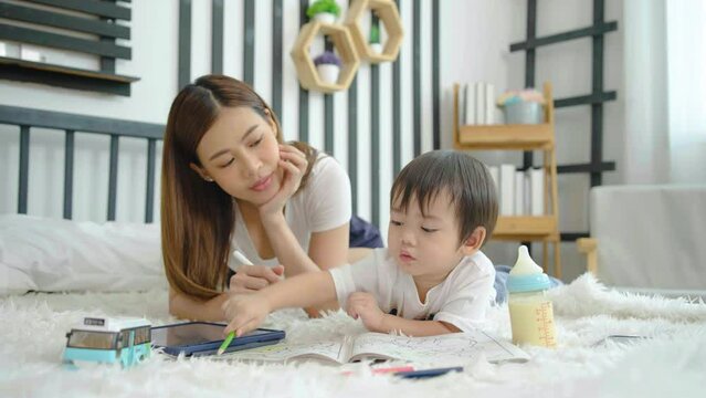 Happy asian little son painting with crayon and colored pencil with his mother in bedroom at home. Mom teaching son how to painting with crayon color on book or doing homework. family psychology