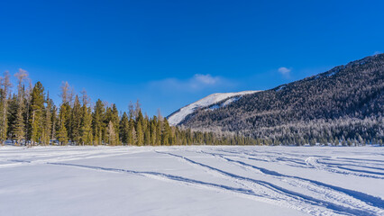 Fototapeta na wymiar In a snow-covered valley, on the surface of a frozen lake, snowmobile tracks are visible. Coniferous forest and mountains against a clear blue sky. Altai. Lower Multinskoe Lake