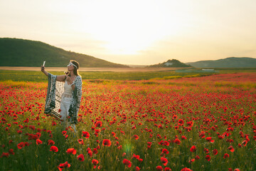 A woman eats strawberries at sunset in a field of poppies and takes selfies on her cell phone....