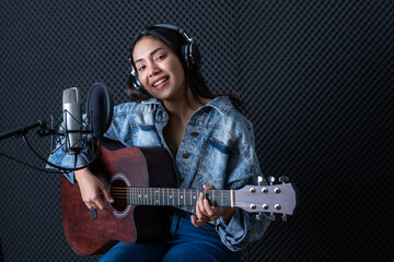 Obraz premium Happy cheerful pretty smiling of portrait a young Asian woman vocalist Wearing Headphones with a Guitar recording a song front of microphone in a professional studio