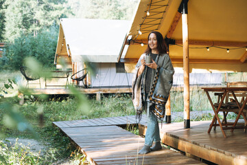 Young brunette woman in poncho drinking tea and relaxing in glamping in nature - obrazy, fototapety, plakaty