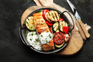 Salmon steak with vegetables and white sauce on dark background.
