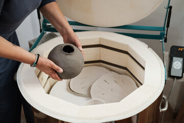 close-up of hands putting vase into the kiln, oven for roasting of unbaked clay handmade products, Unfinished ceramics and utensil. Modern pottery oven ready for firing. - obrazy, fototapety, plakaty