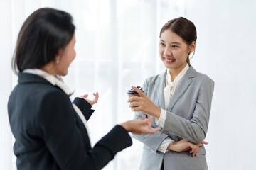 Two Asian business women discuss work with a smile while holding a cup of coffee in the office.