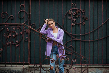 Obraz na płótnie Canvas A portrait of a smiling girl with blond hair, in a purple jacket and a white tank top and jeans, with chic makeup and a stylish hairstyle, stands near a metal fence near the wall of the building.