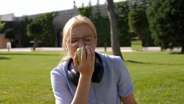 A schoolgirl girl with glasses and headphones eats a green apple in the park.