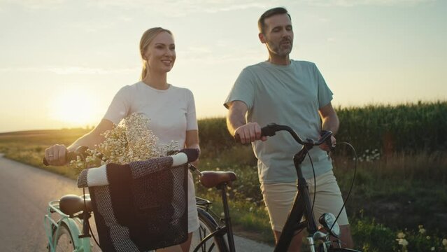 Cheerful caucasian couple of middle age walking with a bike on sunset on village road. Shot with RED helium camera in 8K.   
