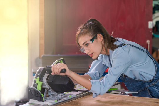 Young Asian Carpenter Woman Using Electric Circular Saw Machine Cutting Wood In Carpentry Wood Working Workshop,  