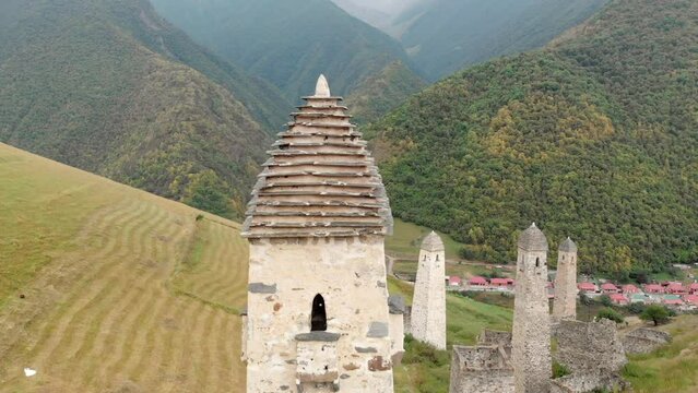 Aerial view of a row of ancient watch towers in a valley among high green mountains. Camera flies up to a nearest stone structure while birds are flying away from its roof