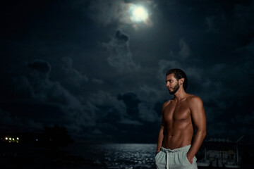 Strong young bearded man on the sea beach at full moon night.