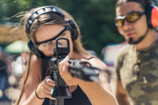 Strong long-haired caucasian girl pursuing her hobby by shooting from a black rifle with collimator. Male instructor in the background. Closeup portrait. Blurred foreground. . High quality photo