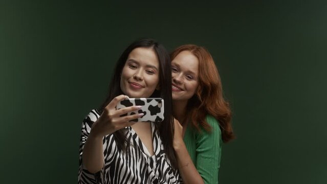 A young asian and young white ginger woman taking a selfie together on a green background 